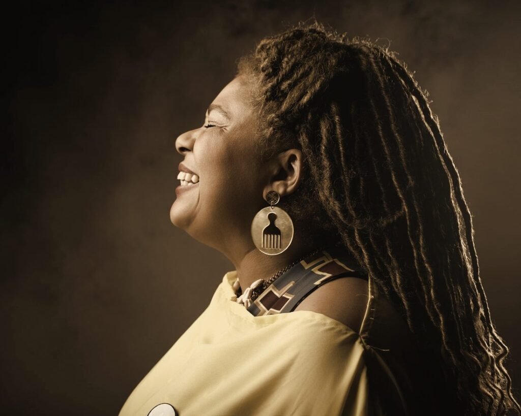 Cristiane Machado de perfil, sorrindo, com tranças longas e brinco dourado, em retrato em tons sépia
