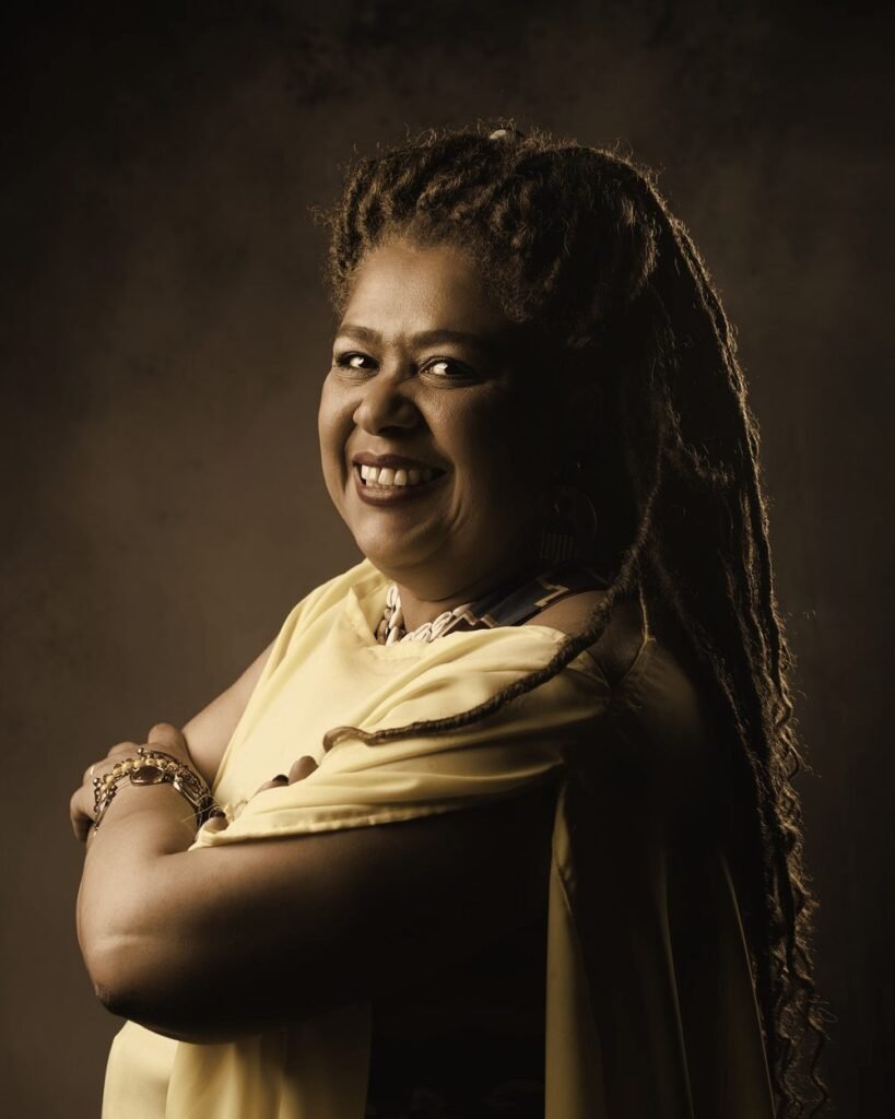 Cristiane Machado sorrindo, de braços cruzados, em retrato em tons sépia sobre fundo escuro