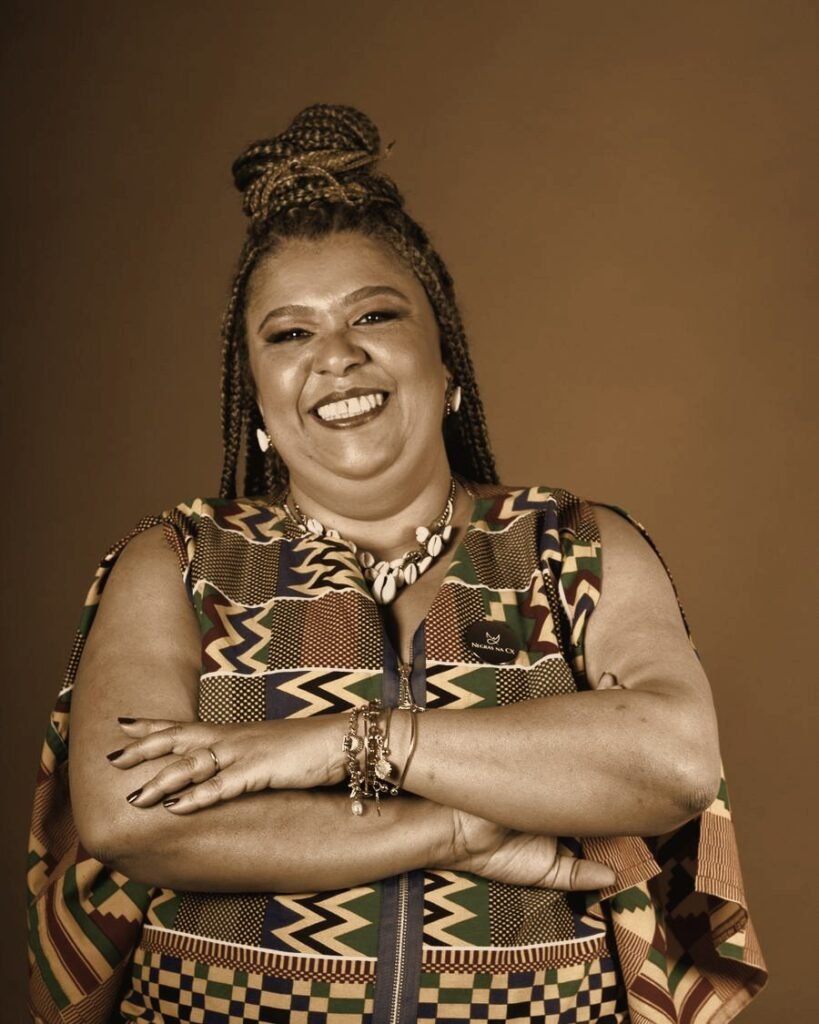 Retrato de Cristiane Machado sorrindo, de braços cruzados, com roupa de estampa africana, em tons quentes